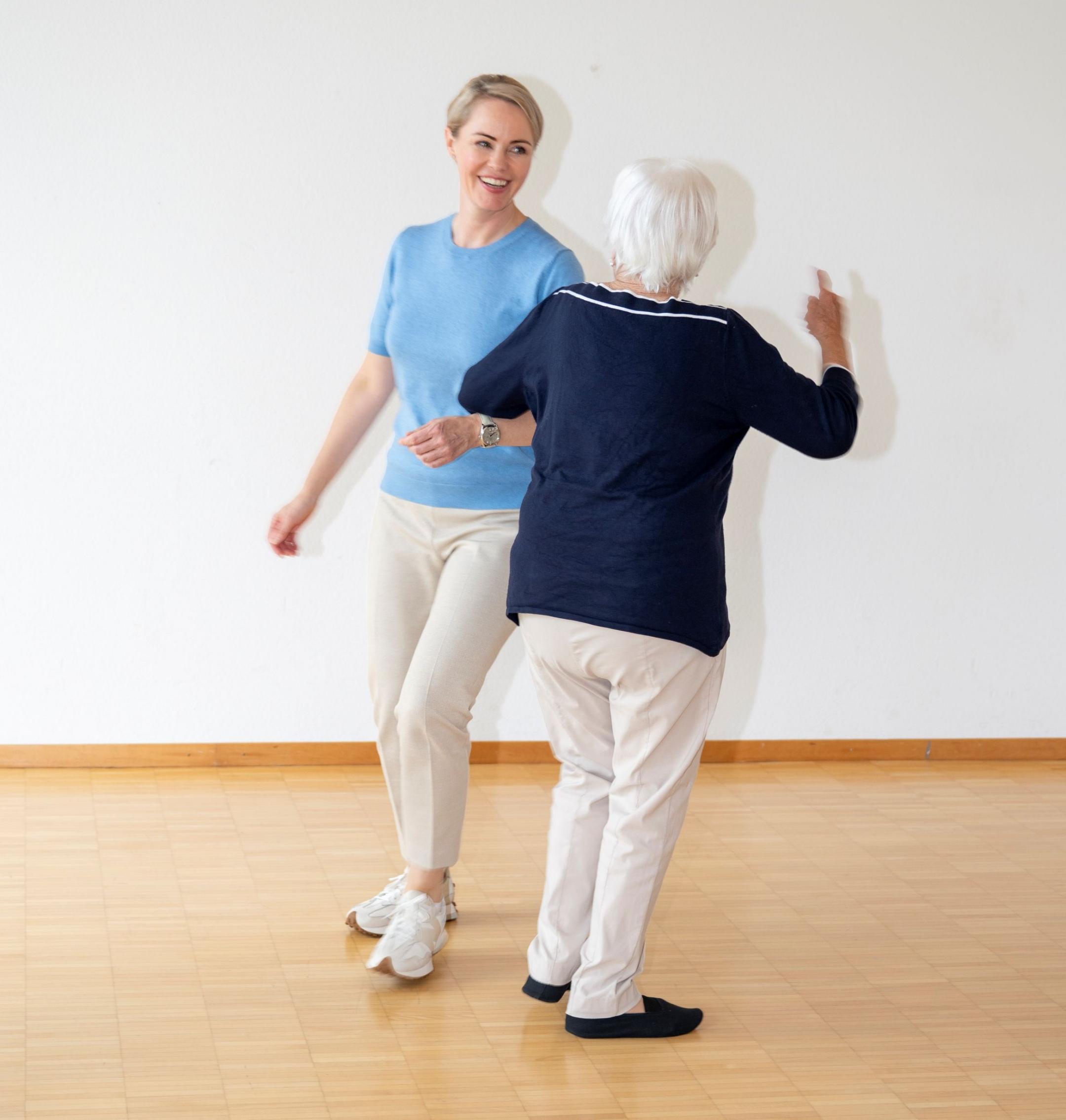 Die lächelnde Gesundheitsdirektorin Natalie Rickli in blauem Oberteil trainiert Tanzschritte mit einer Seniorin in blauem Pullover. Beide tragen helle Hosen.