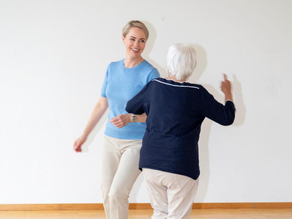 Die lächelnde Gesundheitsdirektorin Natalie Rickli in blauem Oberteil trainiert Tanzschritte mit einer Seniorin in blauem Pullover. Beide tragen helle Hosen.