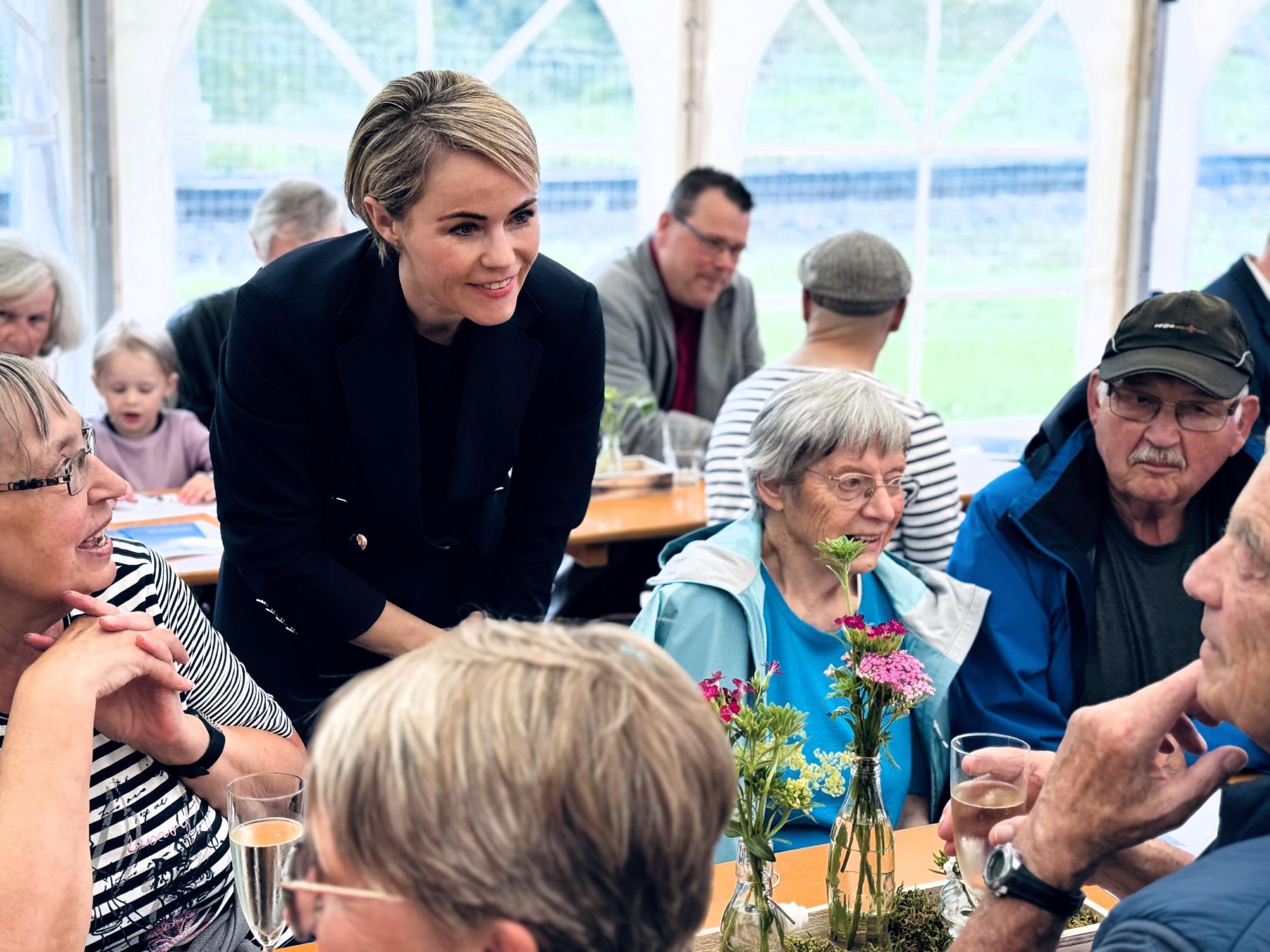 Regierungsrätin Rickli beugt sich lächelnd zu älteren Menschen an einem Tisch im Festzelt, es wird das Jubiläum des Altersheims gefeiert.
