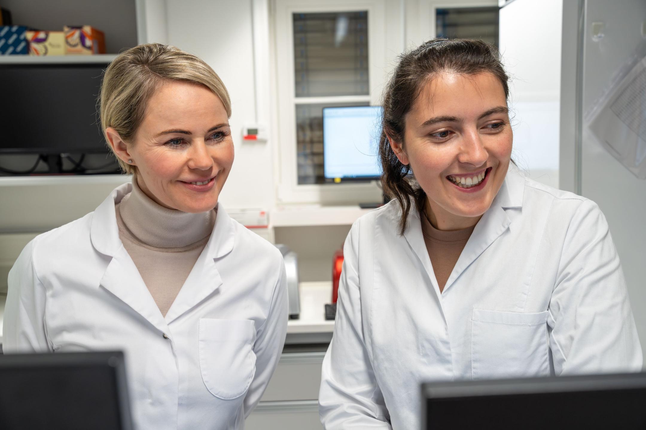 Natalie Rickli und eine Laborantin, beide in weissen Kitteln, schauen lächelnd auf einen Bildschirm.