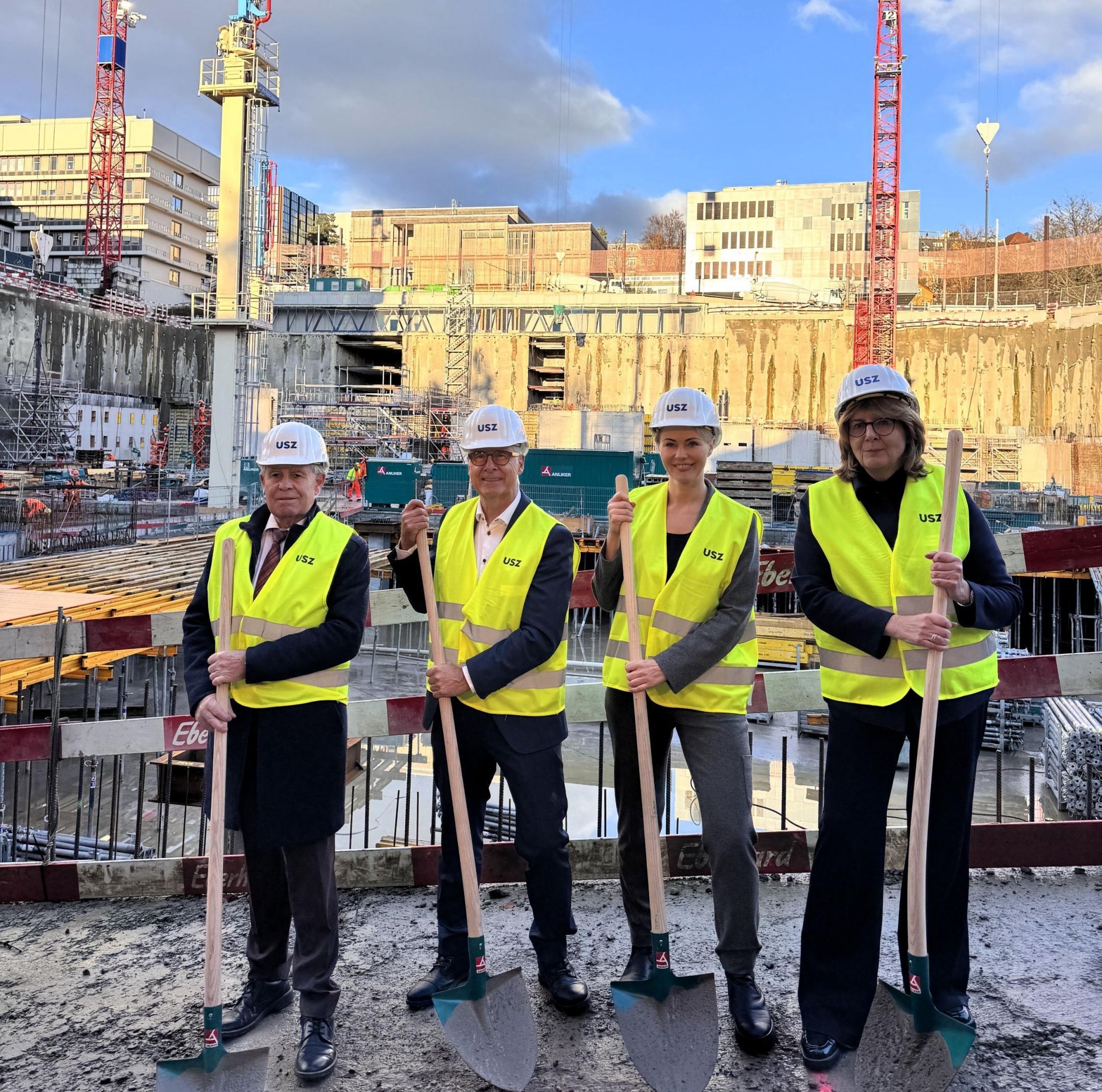Regierungsrat Stocker, Spitalratspräsident Zemp, Regierungsrätin Rickli und CEO Jänicke in gelben Warnwesten und weissen Helmen mit «USZ»-Logo halten Spaten und stehen vor der grossen Baugrube des Universitätsspitals Zürich.