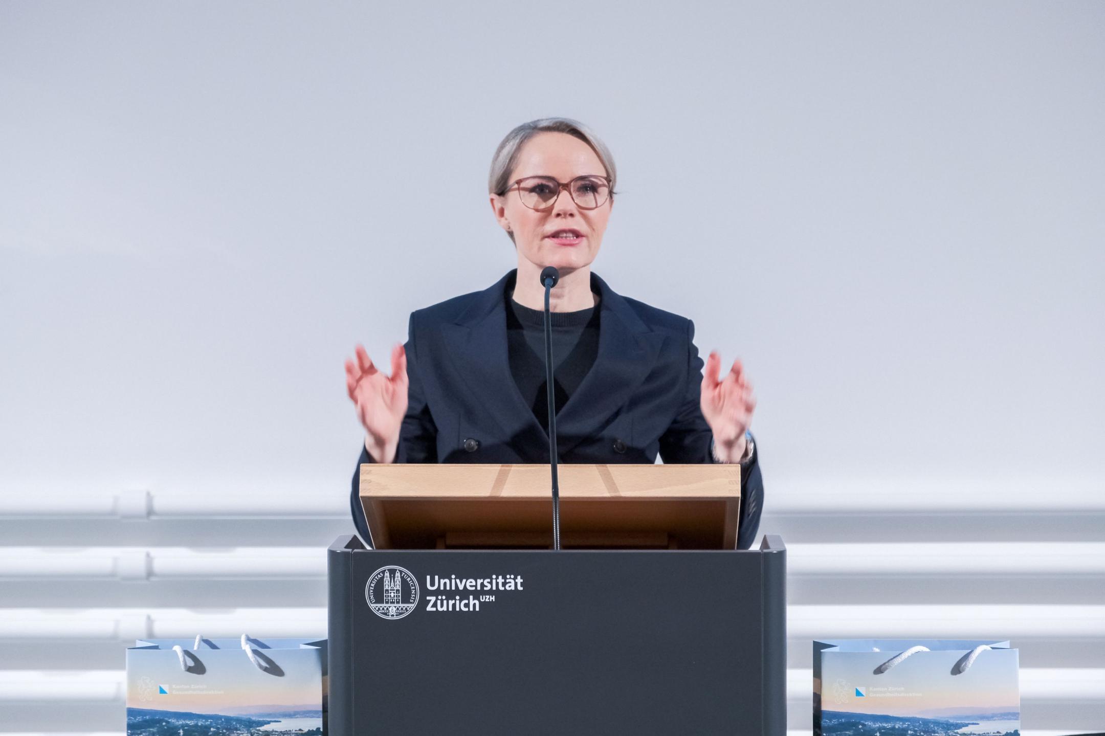 Regierungsrätin Rickli in dunkelblauem Blazer und Pullover sowie Brille tragend und mit Händen referierend am Rednerpult ins Mikrofon sprechend.
