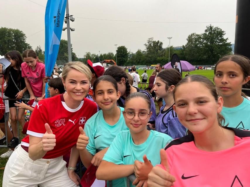 Eine lächelnde Regierungsrätin Rickli in rotem Schweizer Trikot und vier Mädchen zeigen Daumen hoch beim «Schüeli Dietikon», im Hintergrund Fussballplatz und viele weitere Jungen und Mädchen in Sportdresses..