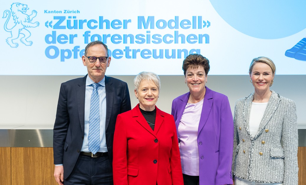 RR Mario Fehr, RR Jacqueline Fehr, RR Silvia Steiner und RR Natalie Rickli stehen lächelnd nebeneinander vor einer Präsentationsfolie zum Zürcher Modell der forensischen Opferbetreuung und blicken in die Kamera