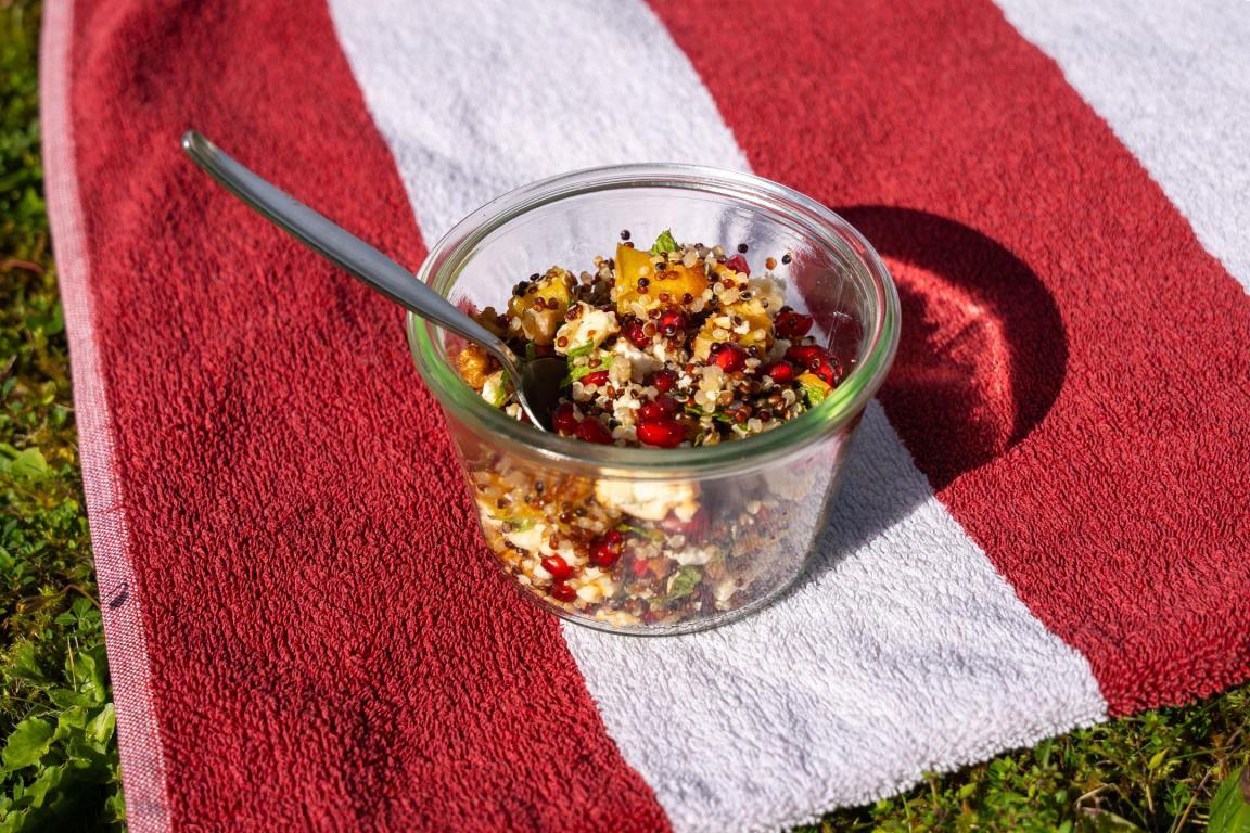 Ein rot-weiss gestreiftes Badetuch auf einer Wiese. Auf dem Tuch steht eine Glasschüssel mit Löffel gefüllt mit Quinoa-Salat.