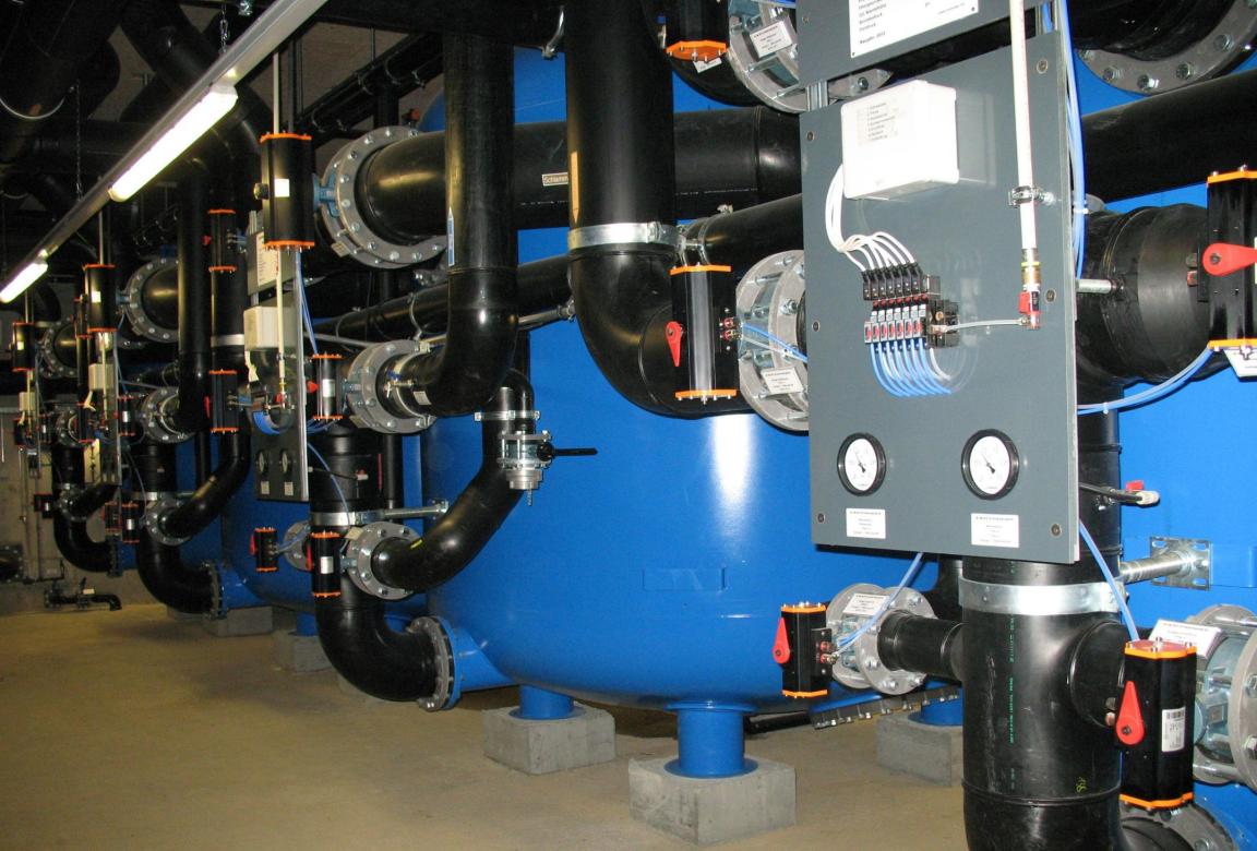 Anlage zur Wasserfilterung mit mehreren blauen Tanks, schwarzen Rohren und einem grauen Kontrollfeld mit drei Manometern..