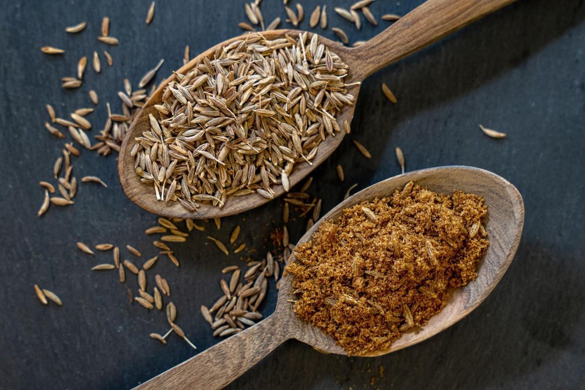 Zwei Holzlöffel mit Kreuzkümmel: ganzer Kreuzkümmel und gemahlener Kreuzkümmel auf dunklem Hintergrund..