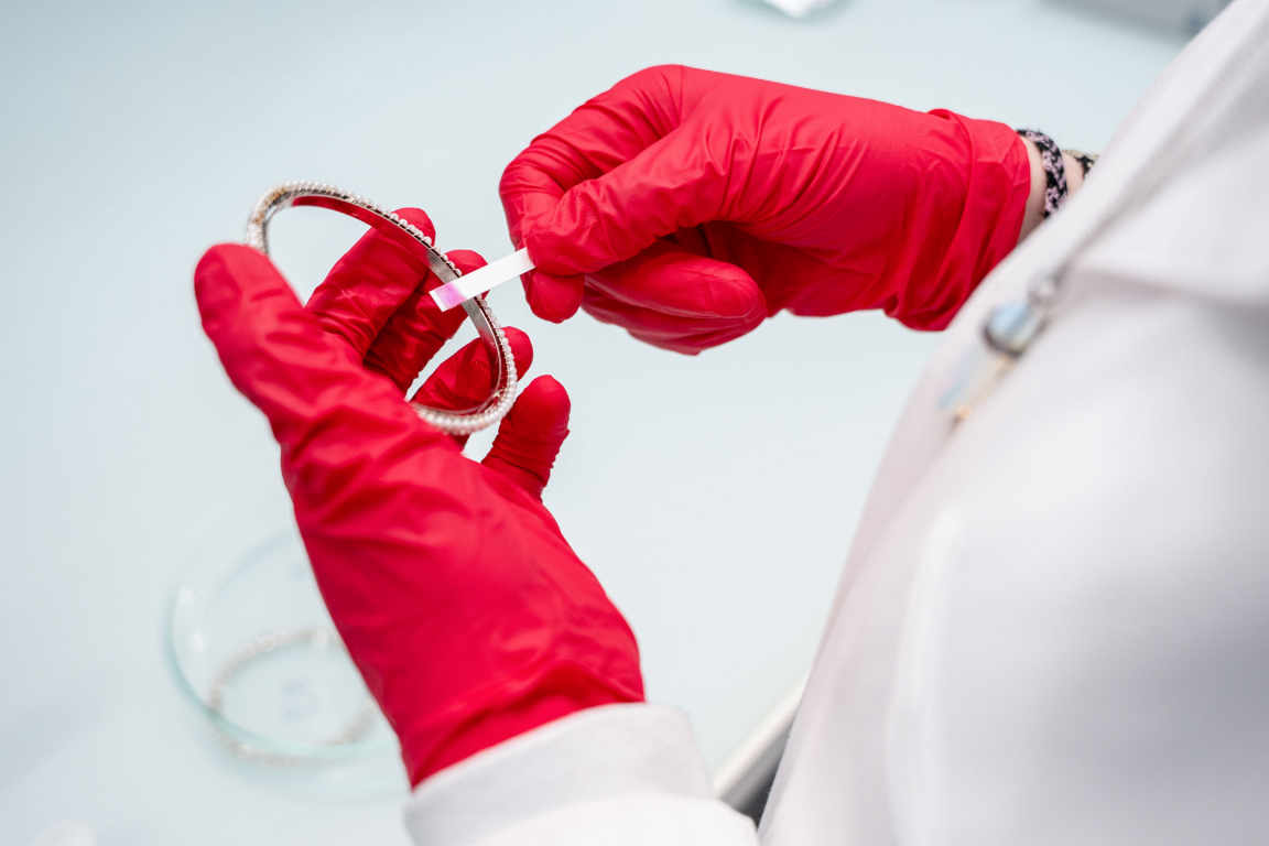 Eine Person mit weissem Labormantel und mit roten Laborhandschuhen hält einen Armreif in der linken Hand. Mit der rechten Hand hält sie einen Teststreifen, der sich pink verfärbt hat. 
