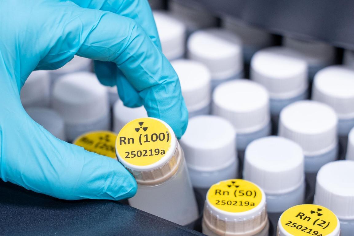 Eine Hand mit einem blauen Schutzhandschuh greift in einem Labor nach einem kleinen Probefläschchen. Das Fläschchen hat ein gelbes Etikett mit einem Radioaktivitäts-Warnsymbol und der Aufschrift «Radon».