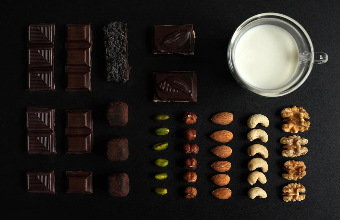 Stücke von dunkler Schokolade und verschiedene Nusssorten sind aufgereiht, oben rechts steht ein Glas mit Milch.