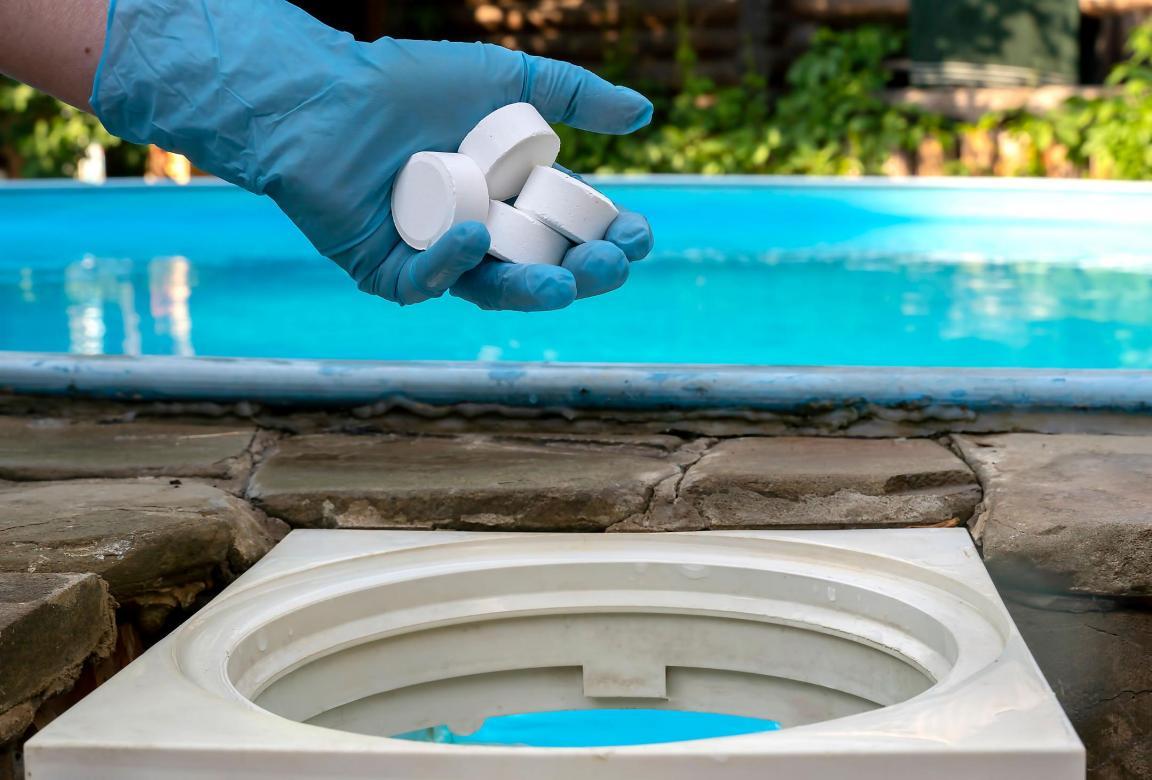 Eine Hand in einem blauen Handschuh hält Chlortabletten vor einem Pool