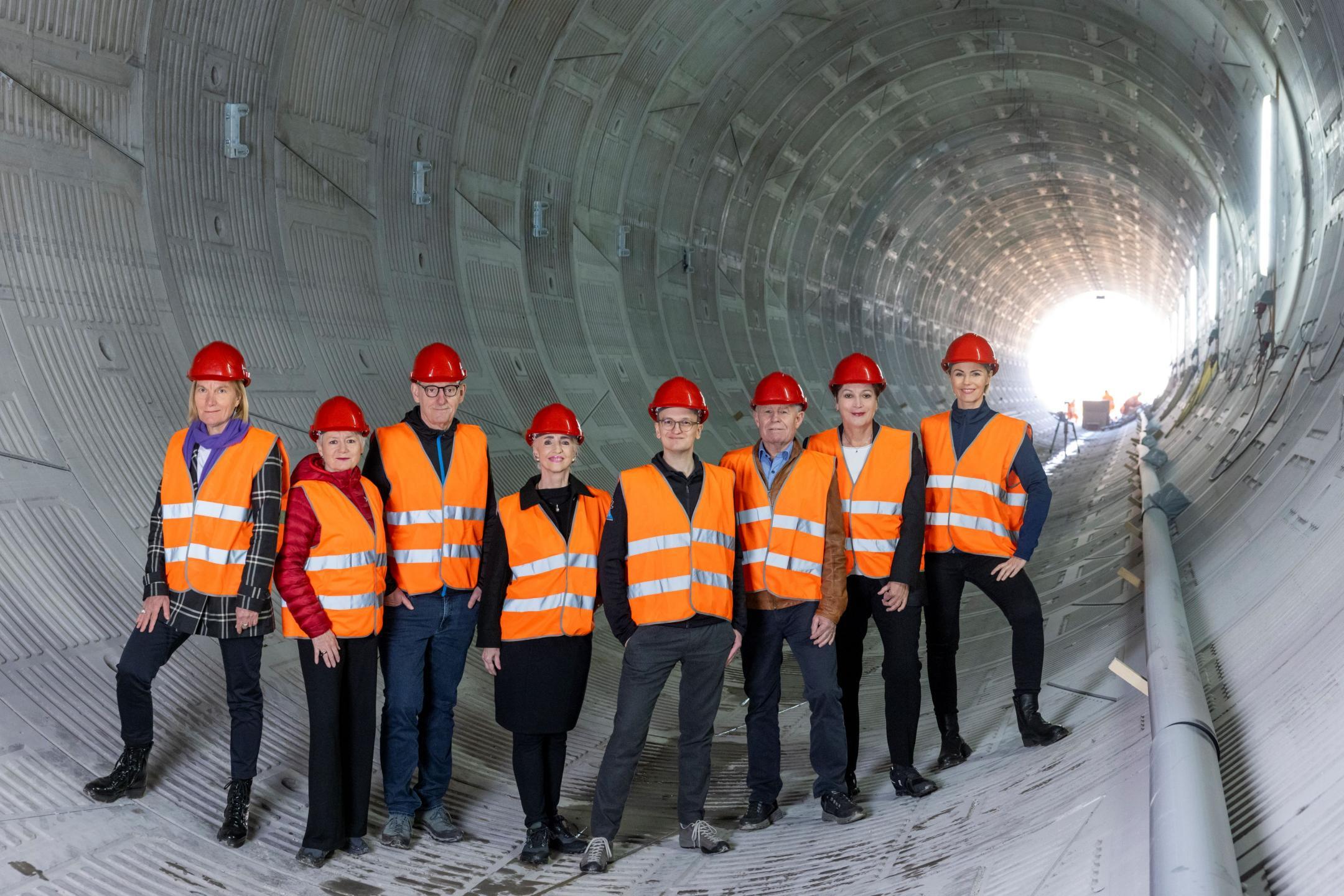 Acht Personen stehen nebeneinander in einem Tunnel, gekleidet in orangen Sicherheitswesten und roten Helmen.