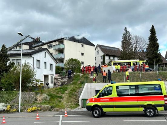 Mehrfamilienhaus bei dem Rauch aus dem Dachstock kommt. Im Vordergrund mehrere Feuerwehrleute.