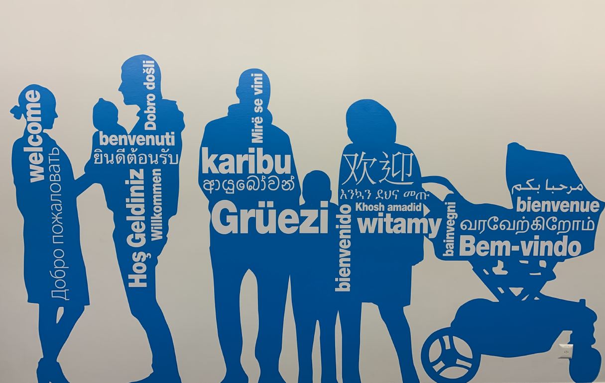 Das Bild zeigt die Wand im Migrationsamt. Darauf sind Personen zu sehen un das Wort Grüezi auf diverse Sprachen. 