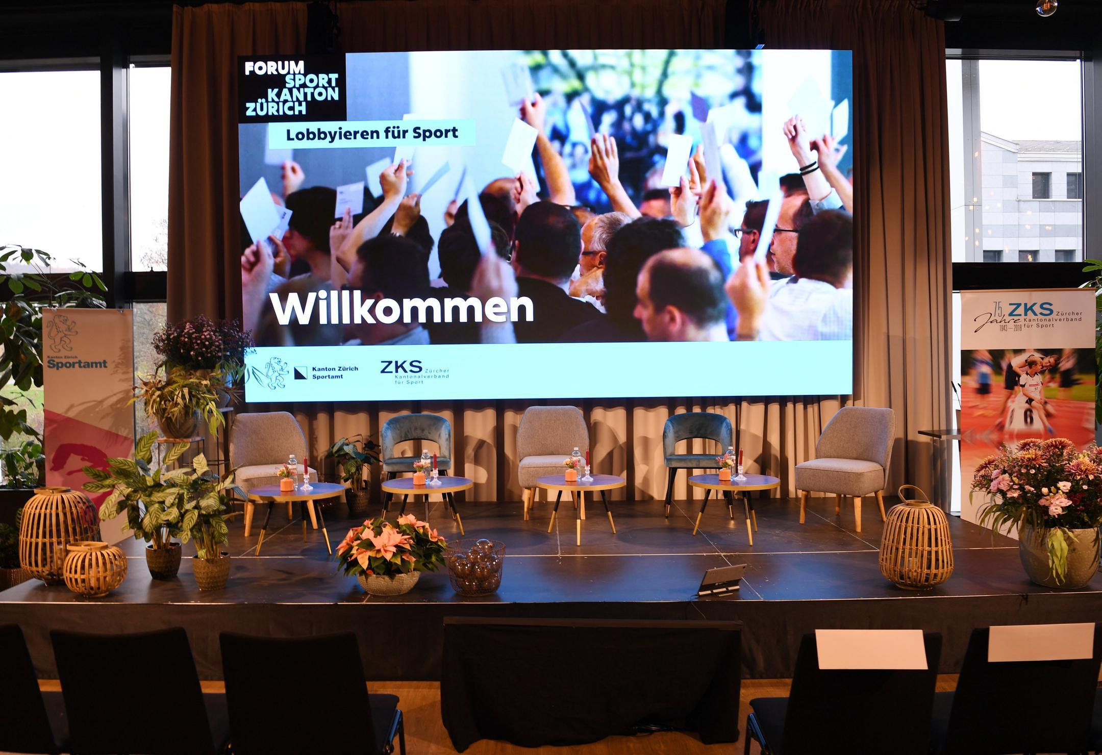 Bühne mit fünf Stuhlen,  vier Beistelltischen und Blumen- und Krezendeko, dahinter eine Leinwand zum Forum Sportkanton Zürich «Willkommen»