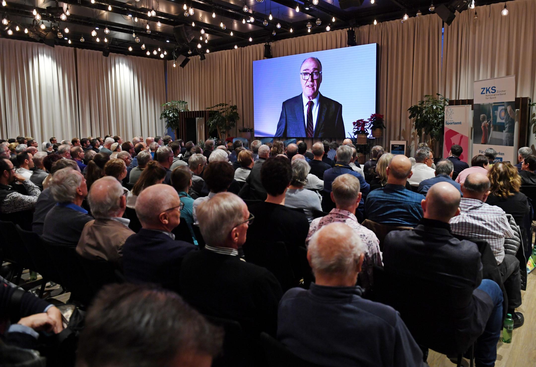Konzertbestuhlung von hinten fotografiert, Teilnehmende des Forums sitzen auf Stühlen, auf der Leinwand vorne wird eine Videobotschaft von Bundesrat Martin Pfister gezeigt