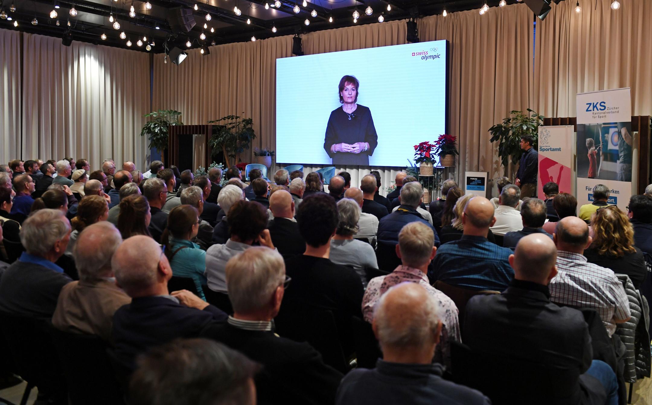 Publikum in Konzertbestuhlung, von hinten fotografiert, auf der Leinwand vorne spricht Rutz Metzler-Arnold in einer Videobotschaft