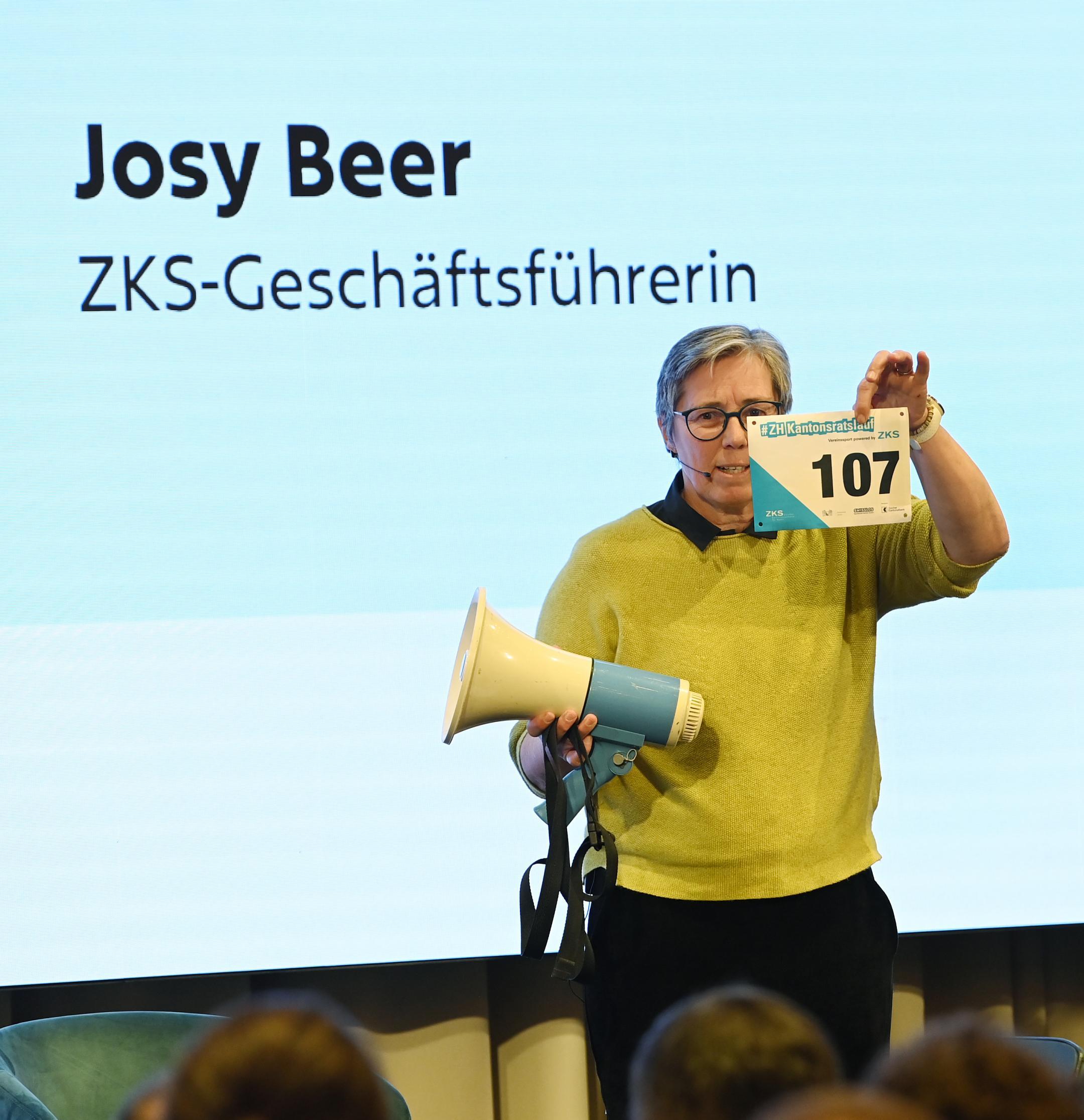eine Frau steht mit einem Megafon und einer Startnummer in den Händen auf der Bühne