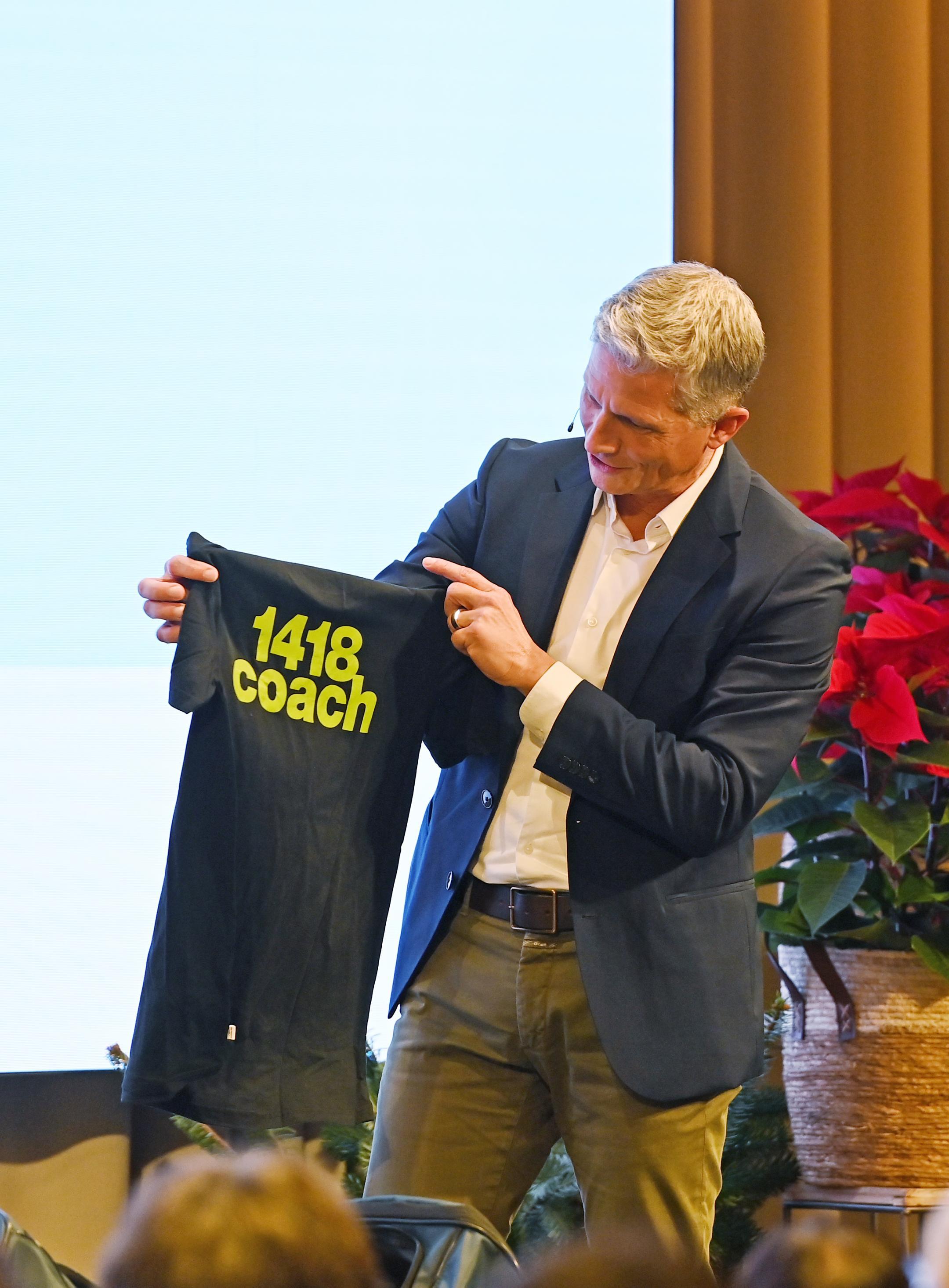 ein Mann steht auf einer Bühne und hält ein T-Shirt mit Aufschrift 1418coach in den Händen