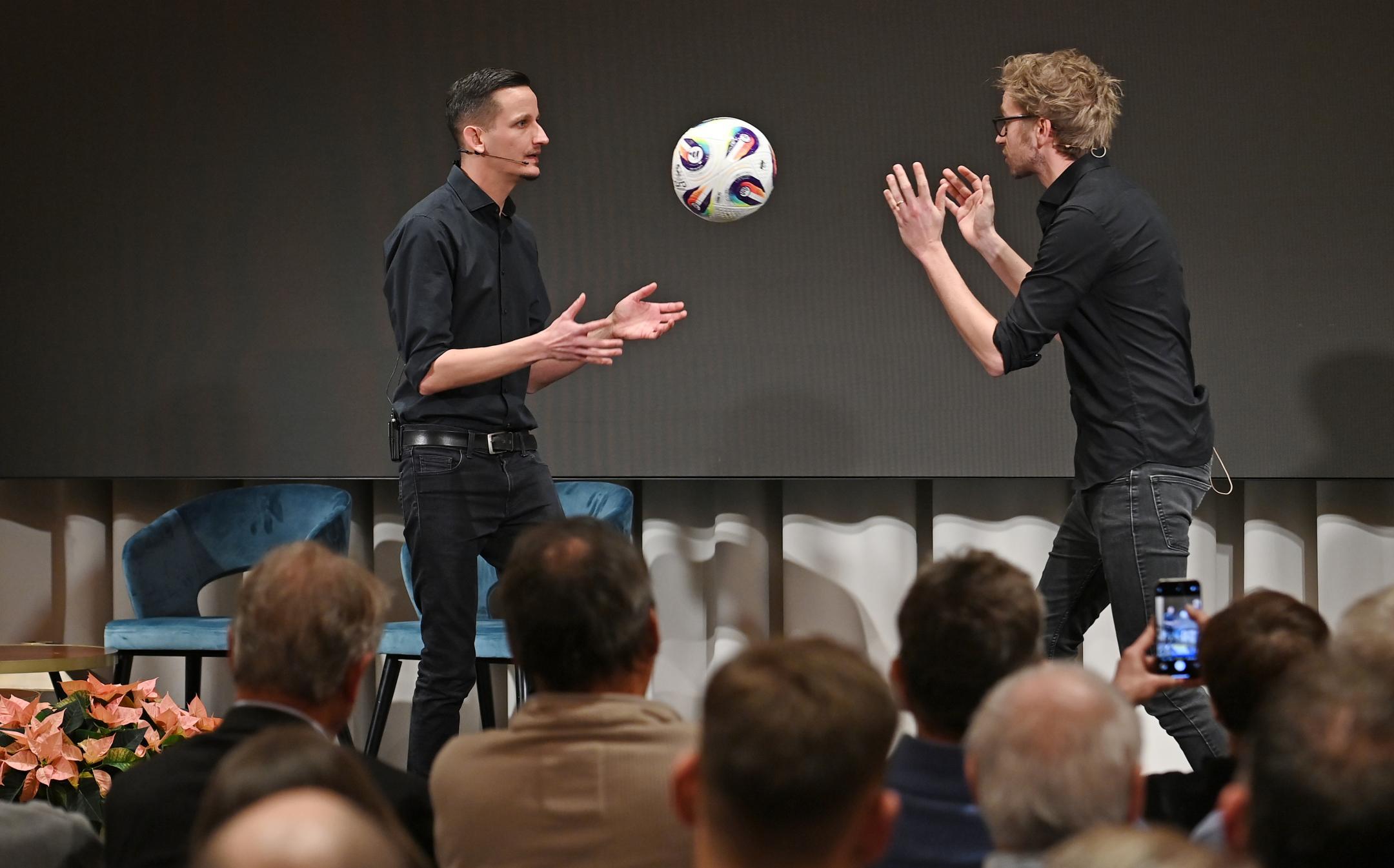 zwei Männer stehen auf der Bühne und werfen einen Fussball hin und her