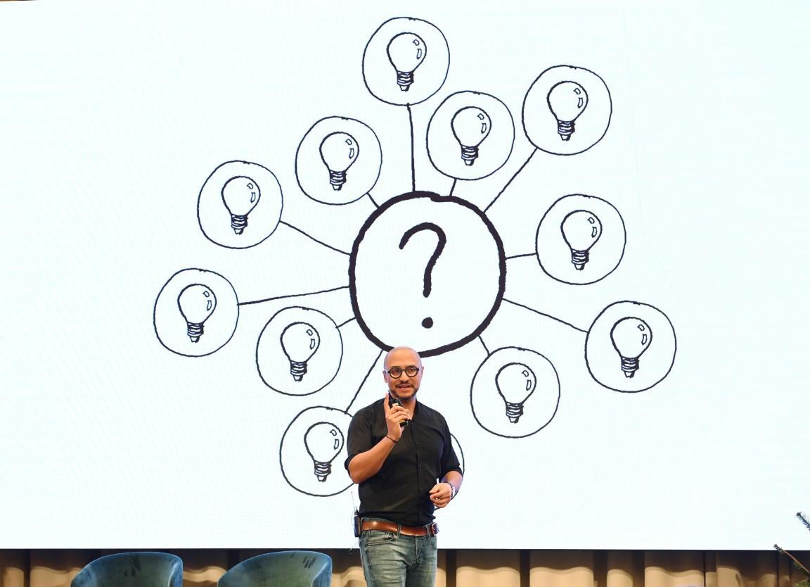 Referent hält einen Vortrag, im Hintergrund ist ine Folie eingeblendet, auf welcher ein Fragezeichen in der Mitte und darum mehrere Glühbirnen gezeichnet sind