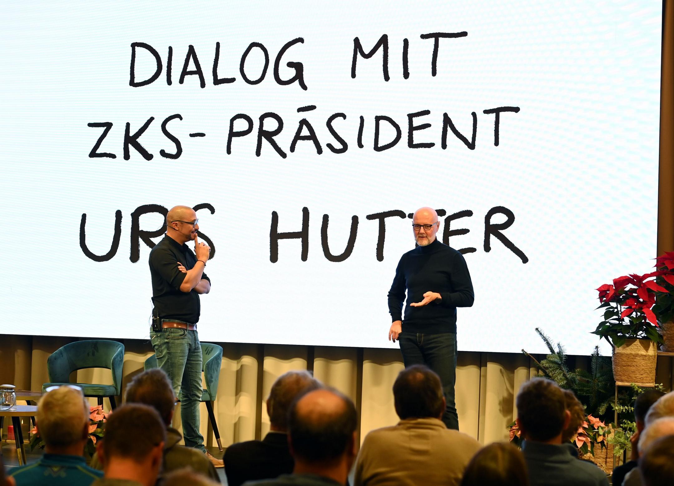 Zwei Männer stehen auf der Bühne und sprechen zusammen, im Hintergrund steht « Dialog mit ZKS-Präsident Urs Hutter» auf der Folie