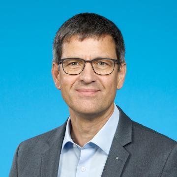 Porträt von Markus Traber, Amtschef, der sich für nachhaltige Mobilität im Kanton Zürich engagiert. Er trägt Brille, hellblaues Hemd und graues Sakko vor blauem Hintergrund.