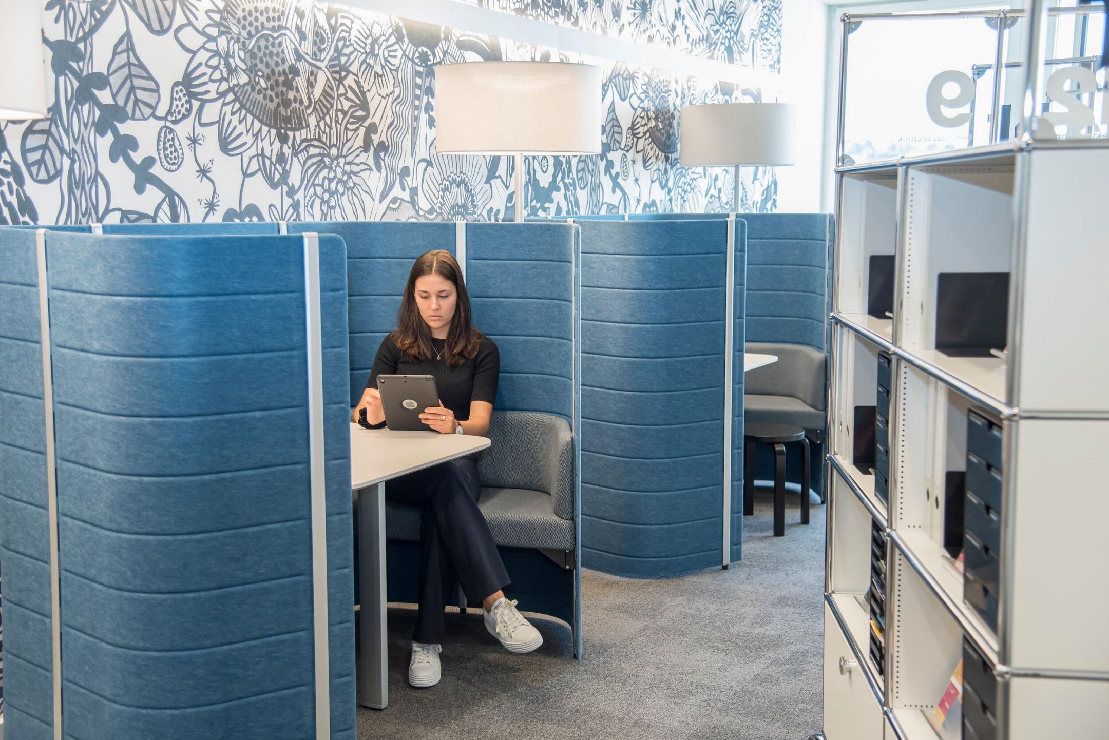 Junge Frau sitzt mit Tablet in einer abgeschirmten blauen Besprechungsnische