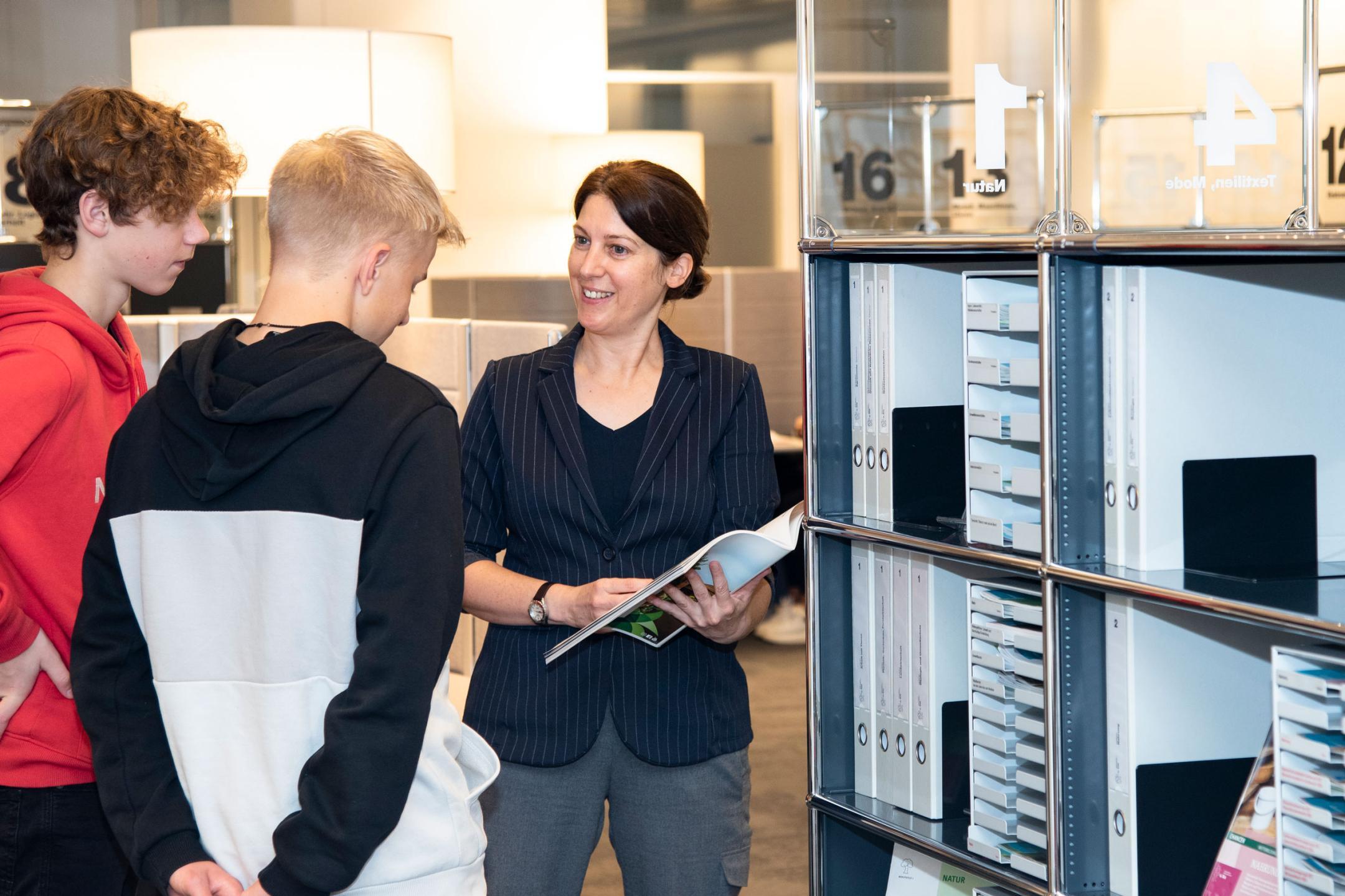 Eine Berufsberaterin sieht sich mit zwei Jugendlichen vor einem Regal in der Infothek eine Broschüre an.