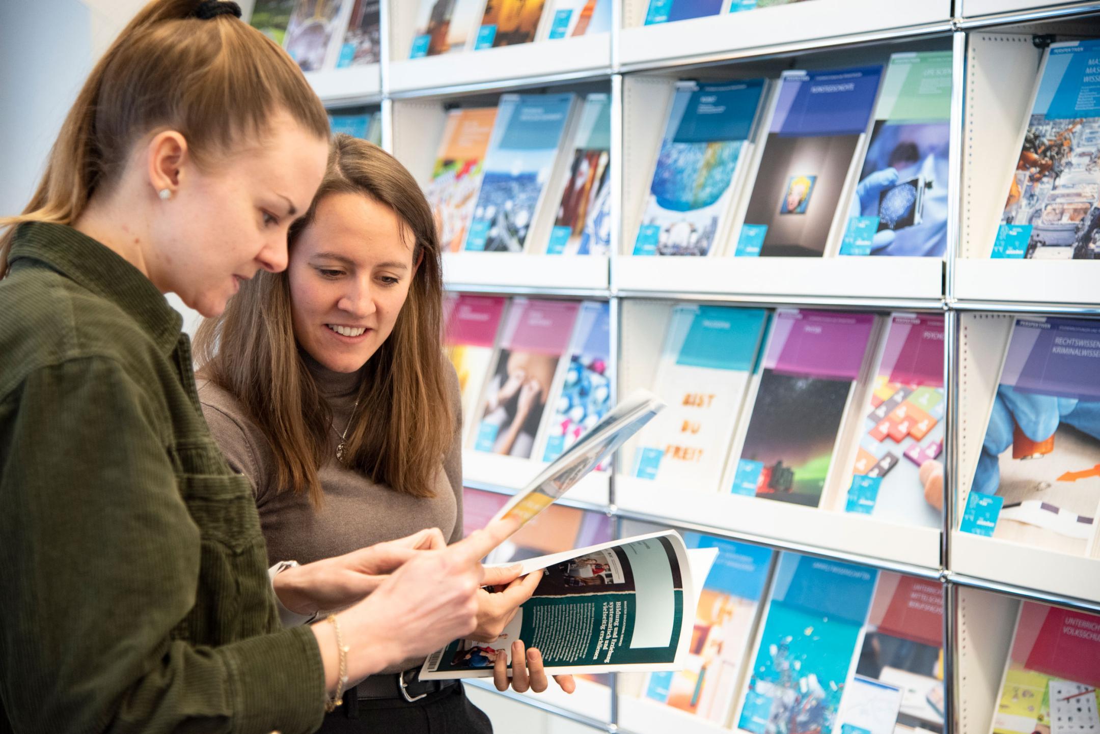 Eine junge Frau betrachtet mit einer Berufsberaterin eine Broschüre vor einem Regal in der Infothek.