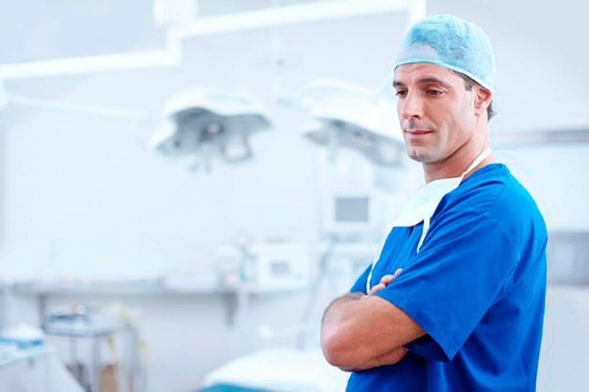 Ein Chirurg steht, etwas nachdenklich, mit verschränkten Armen und abgelegtem Mundschutz in einem Operationssaal.