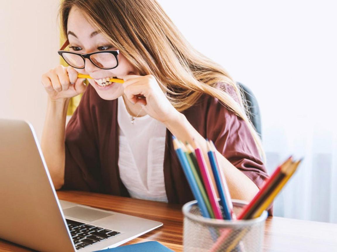 Eine junge Frau mit Brille und langem Haar starrt in einen PC und beisst dabei fest in einen Bleistift.