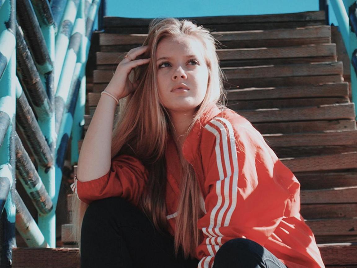 Eine junge blonde Frau mit roter Jacke sitzt auf einer Treppe und scheint in die Ferne zu schauen.