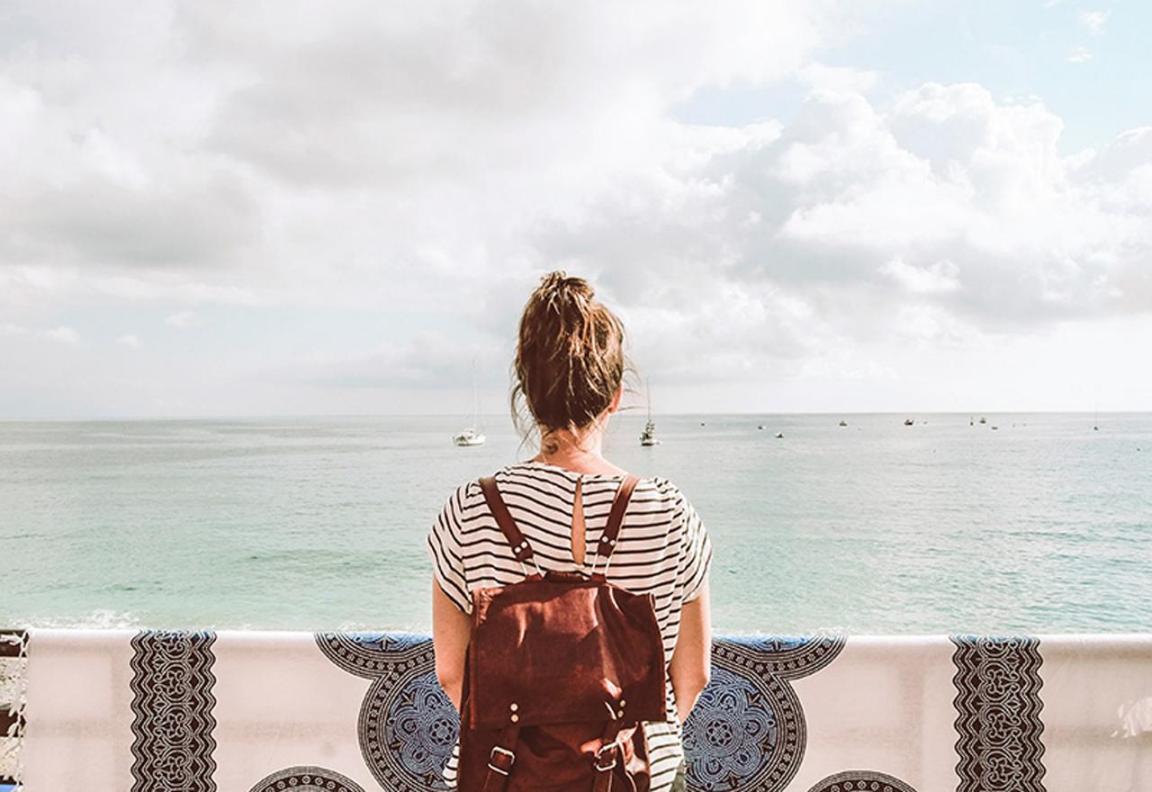 Eine junge Frau mit einem Rucksack, den Rücken zur Kamera gewandt, schaut aufs Meer hinaus.