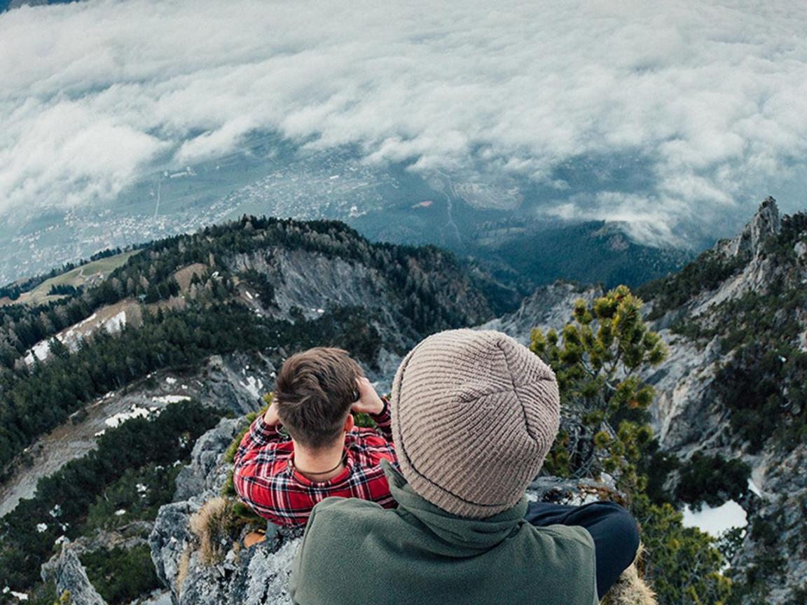 Zwei Personen, mit dem Rücken zur Kamera, stehen in luftiger Höhe auf einem Felsen und schauen hinab ins weit darunter liegende Tal.