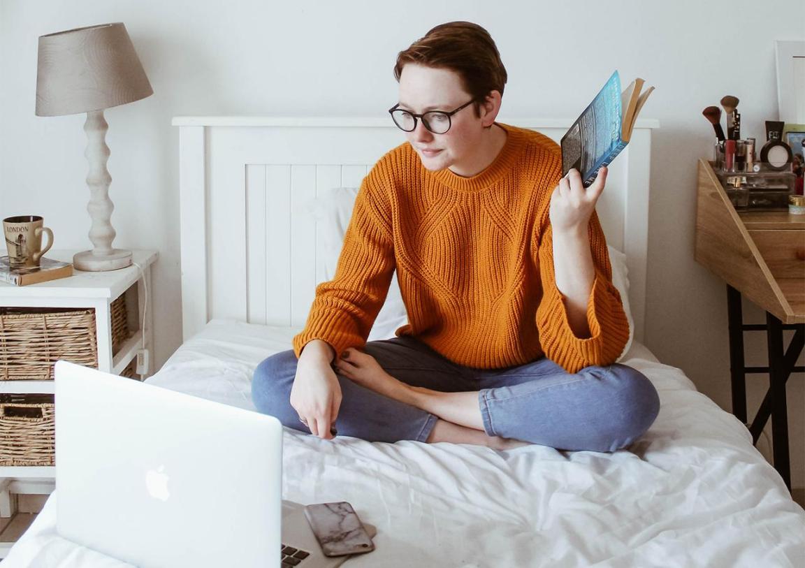 Eine junge Frau mit Brille sitzt auf einem Bett und schaut in einen Mac, in der linken Hand ein Buch.