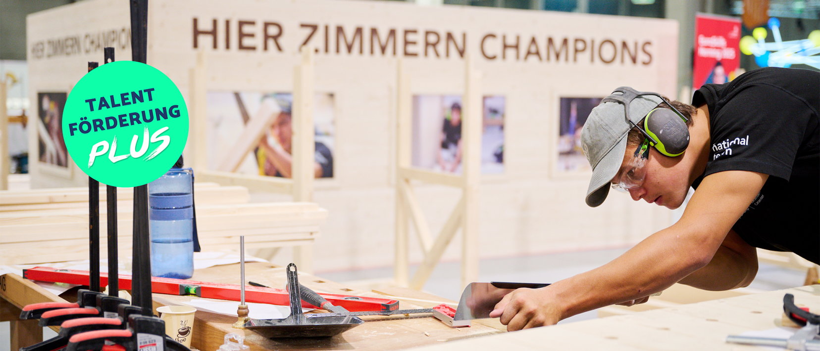 Junger Schreiner mit Kappe, Schutzbrille und Gehörschutz bearbeitet ein Holzstück. Im Hintergrund ein Schriftzug «HIER ZIMMERN CHAMPIONS» und ein grüner Kreis mit «TALENTFÖRDERUNG PLUS».
