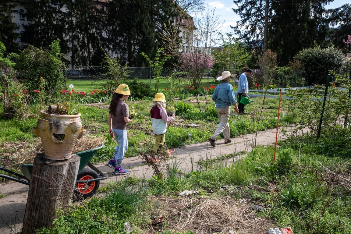 Zwei Kinder und zwei Erwachsene bewegen sich auf einem Weg durch einen grünen Garten mit Beeten, Blumen und Bäumen. Eine Person trägt eine Giesskanne.