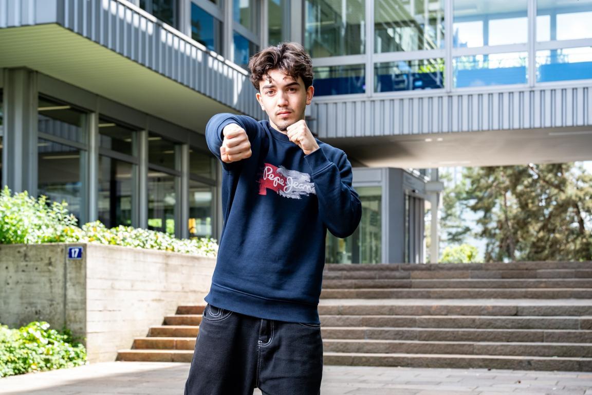 Ein junger Mann in Jeans zeigt eine spielerische Kampfpose vor der Treppe zu seiner Schule. 
