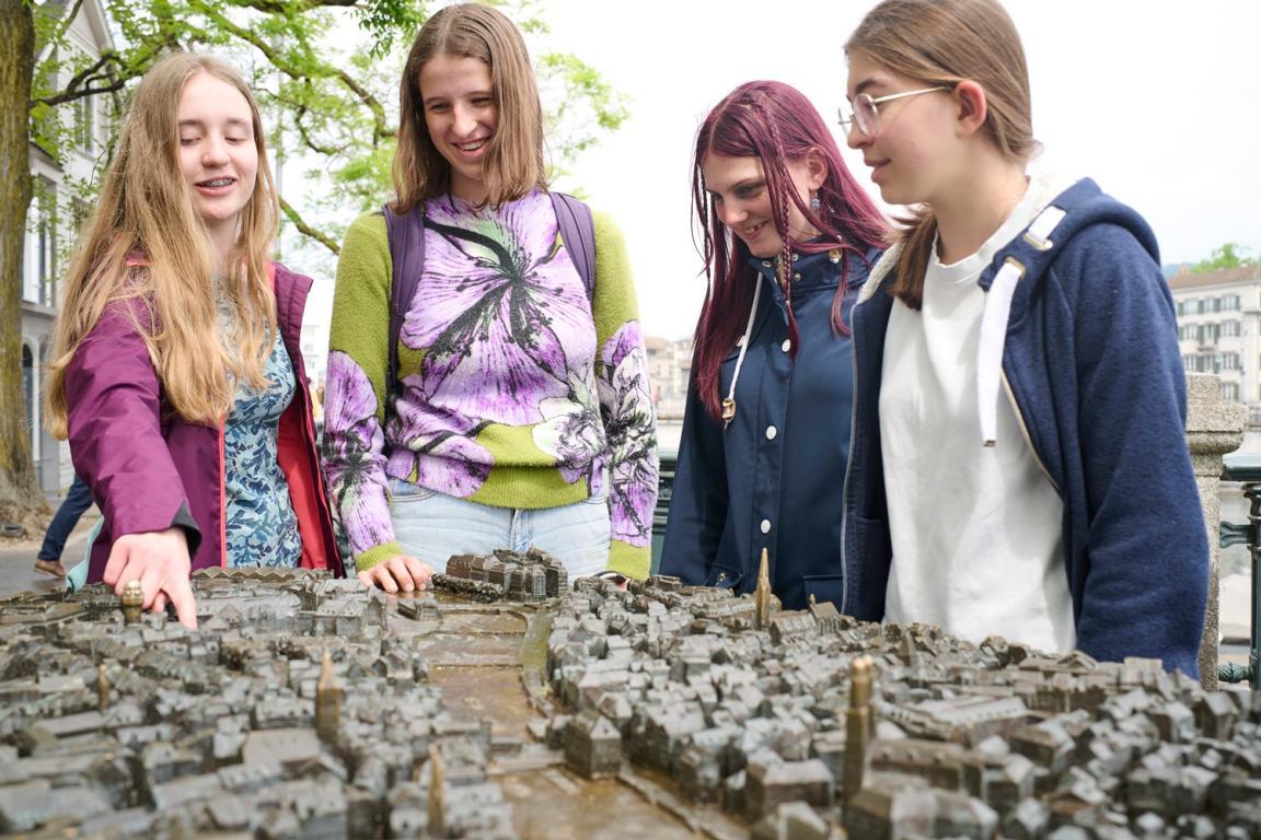 Vier Gymnasiastinnen stehen draussen um ein Modell der Stadt Zürich herum . Ein Mädchen zeigt auf ein Haus des Modells. 