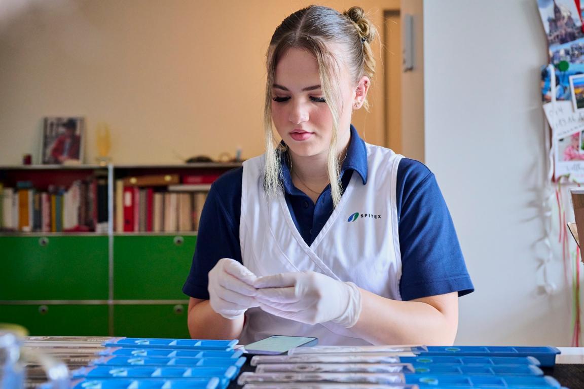 Eine junge Frau sitzt in Arbeitskleidung und mit Handschuehen an einem Tisch und füllt Medikamente ab.