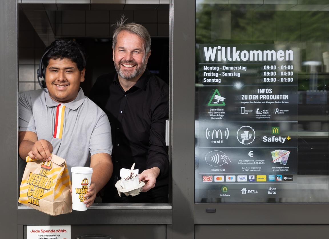 Der Lernende und sein Berufsbildner schauen fröhlich aus dem Fenster des McDonalds Drive-In.