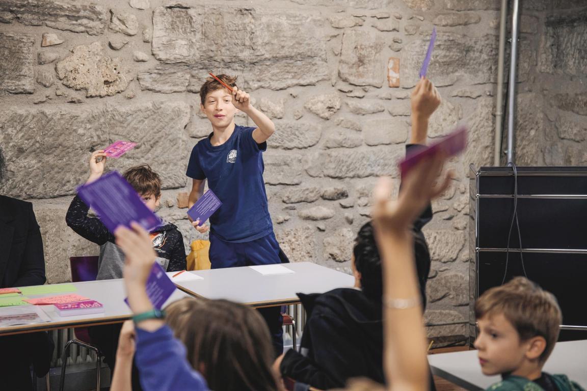 Die Kinder halten violette Karten in die Luft. Ein Schüler steht hinter seinem Tisch und zählt die Zettel. 