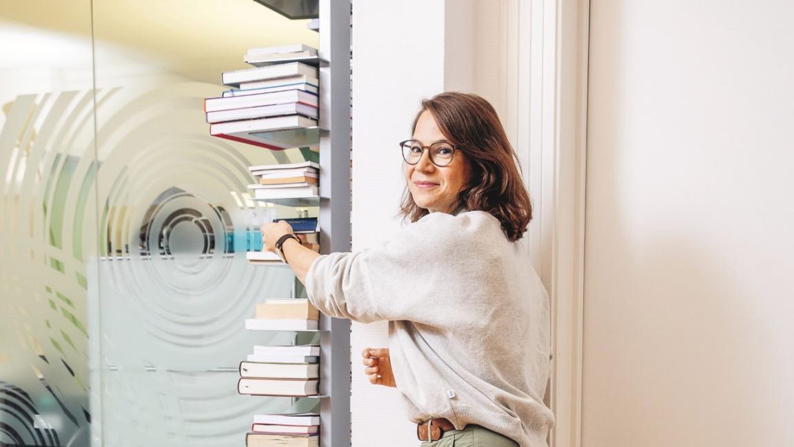 Junge Frau mit Brille schaut, halb abgedreht, in die Kamera und greift nach einem Buch im Regal