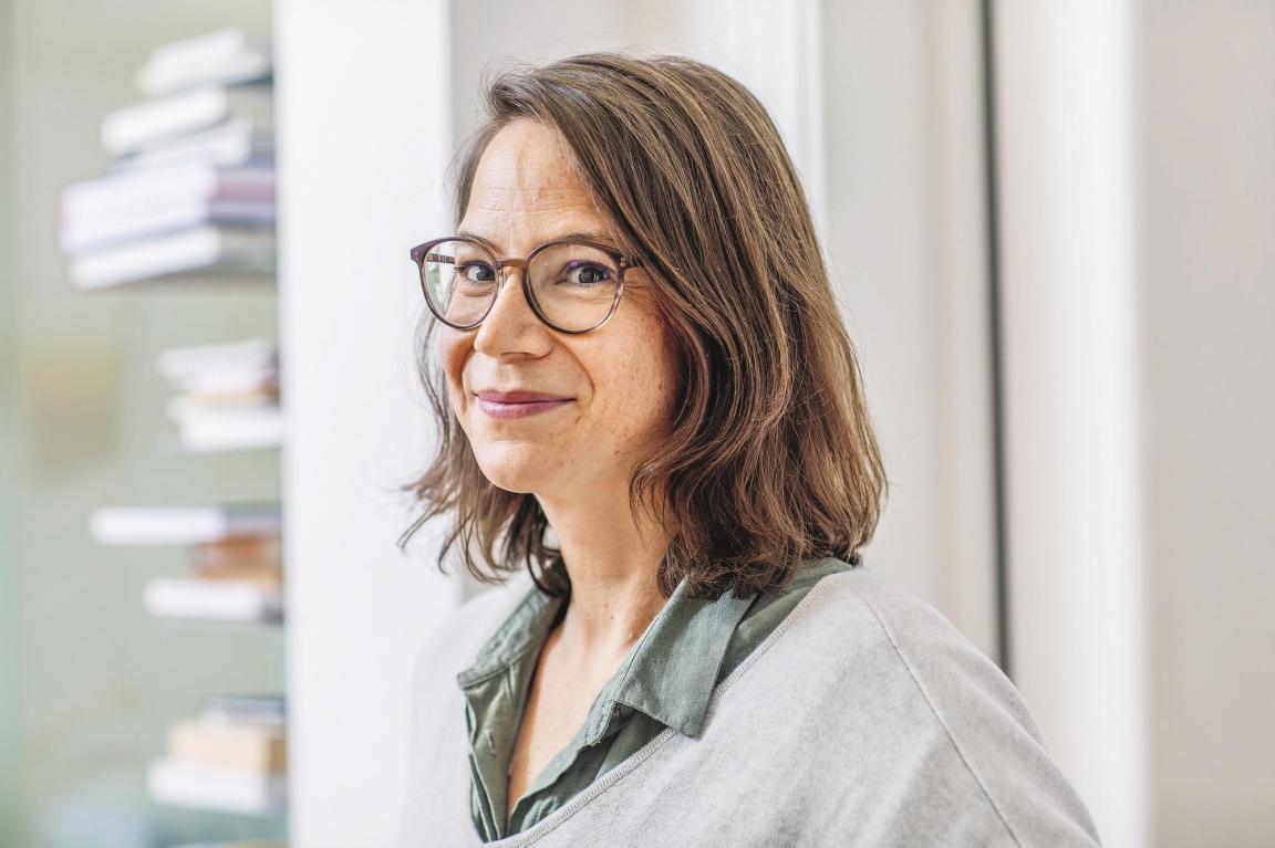 Frau mit Brille und kurzen braunen Haaren lächelt in die Kamera.  Im Hintergrund sind Bücher an der Wand aufgreit.