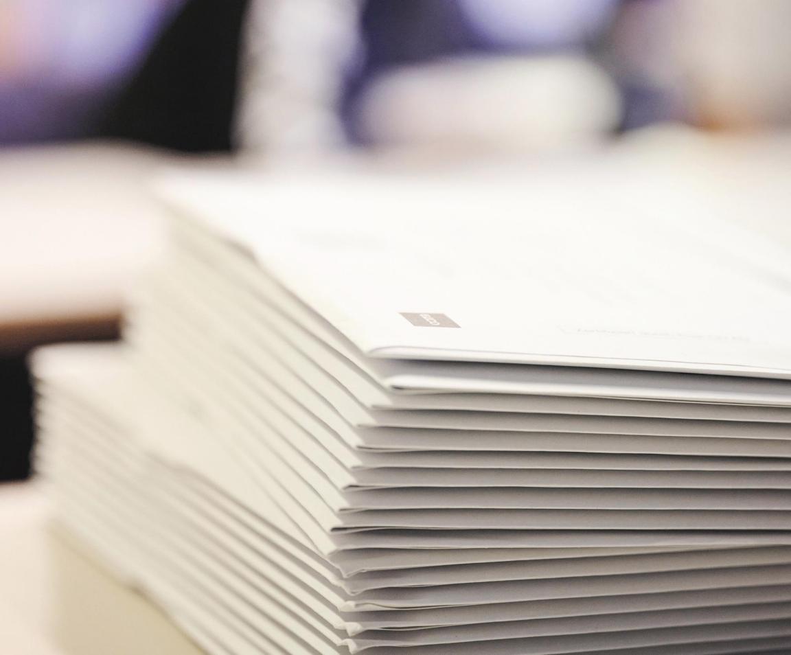 Ein Stapel Abstimmungscouverts liegen auf einem Tisch. Der Hintergrund ist verschwommen, man erkennt den Umriss eines Schülers.