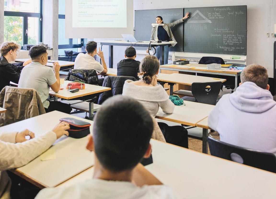 Die Lehrerin steht vor der Klasse und zeigt auf eine Notiz auf der Wandtafel.