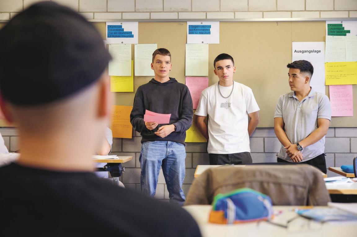 Drei Jugendliche stehen vor einer Pinnwand und präsentieren etwas der Klasse.