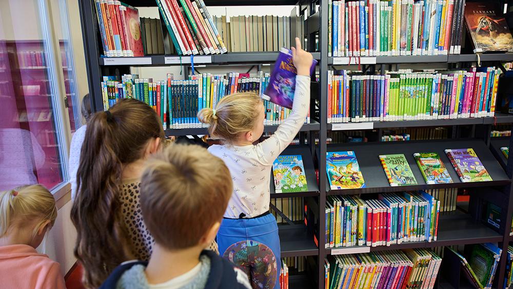 Die Kinder stehen in einer Schlange vor den Bücherregal. Ein Kind holt daraus ein Buch hervor.