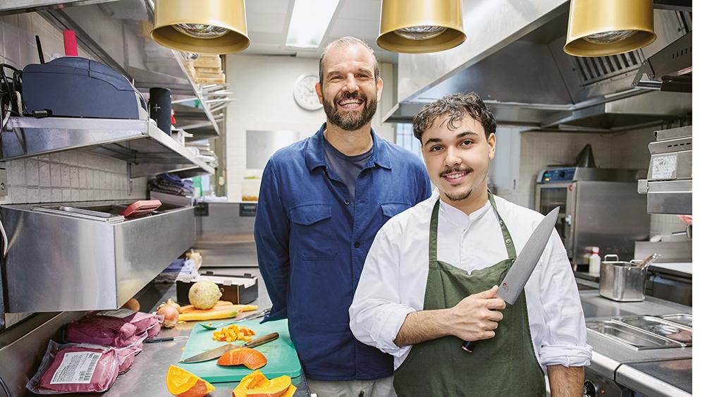 Der Ausbildner steht mit dem Lernenden in  der Küche eines Restaurants. Der Lernende hält ein Messer in der Hand. Auf der Ablage liegt verpacktes Fleisch und geschnittener Kürbis. 